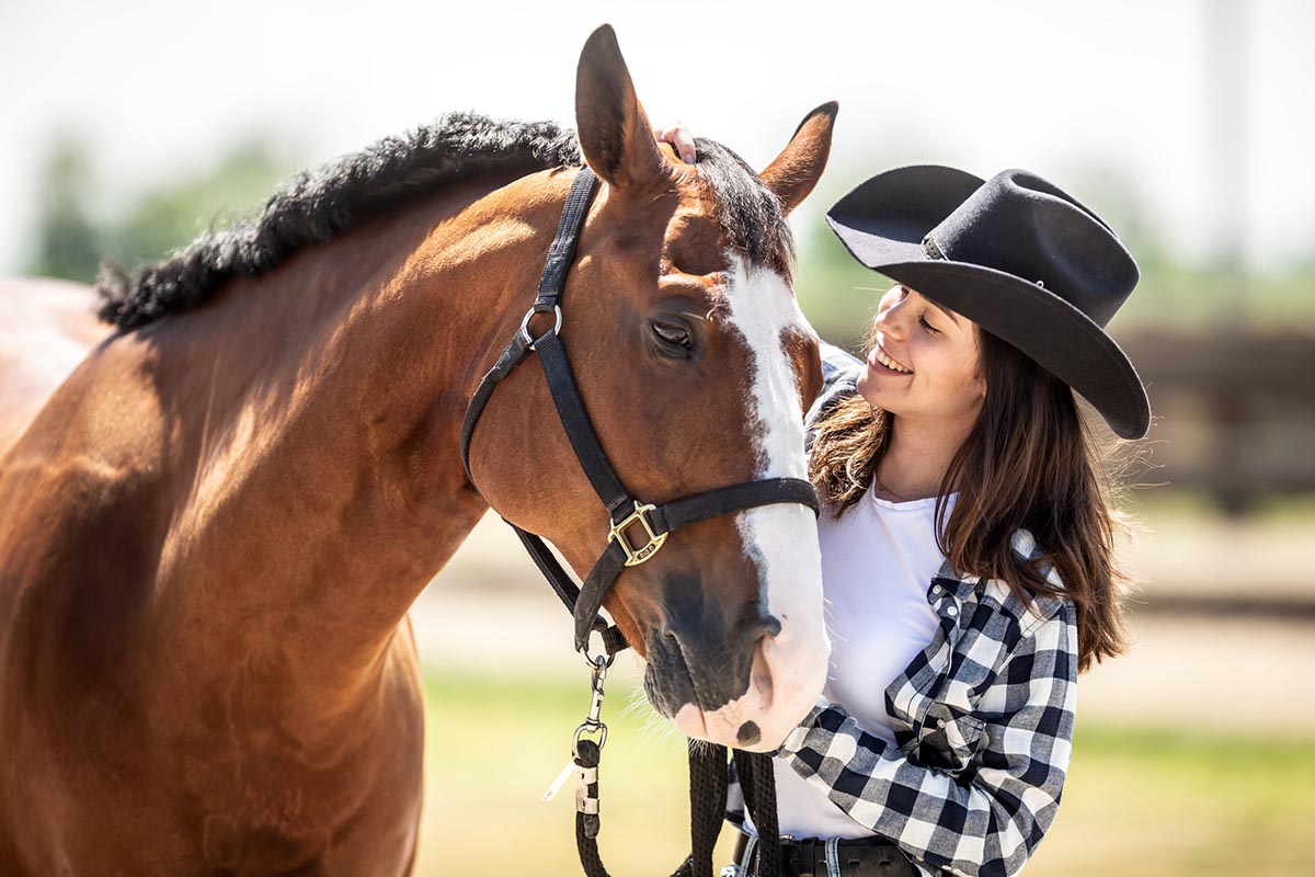 A woman in a cowboy hat and plaid shirt smiles and pets her bay horse