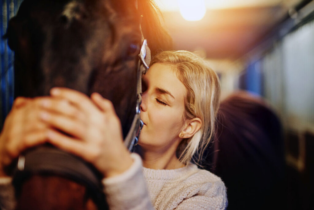 a young blond woman hugs and kisses her bay horse's face and neck