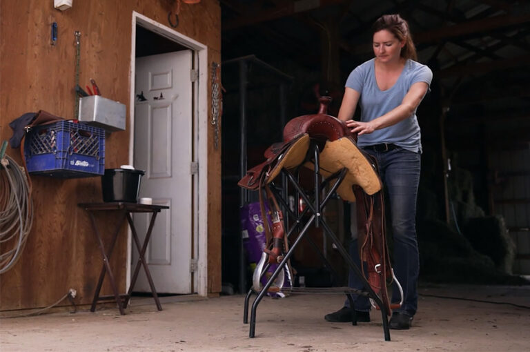 a woman cleans a leather western saddle in a barn aisle