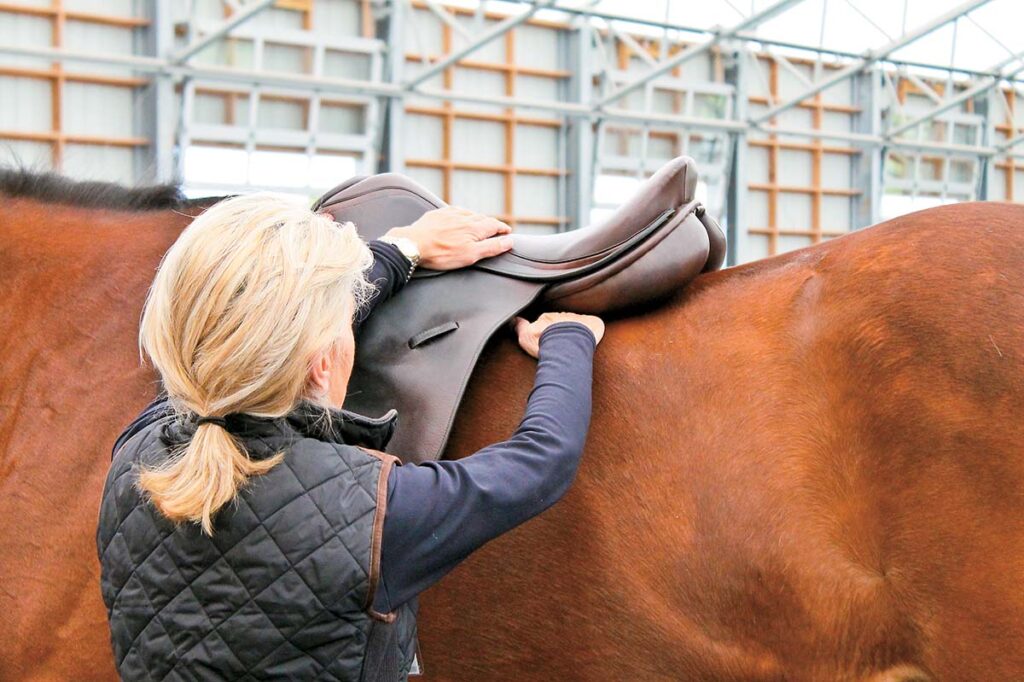 A blond woman runs her hand underneath an English saddle placed on a bay horse's back to assess whether it fits properly