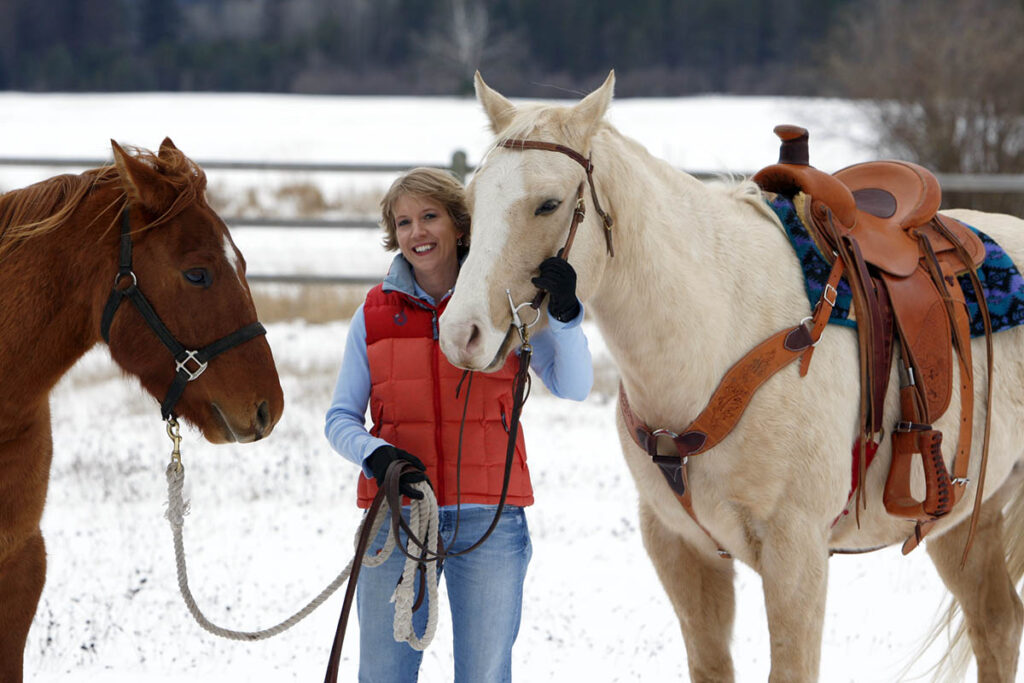 A middle aged woman wearing a red vest leads two horses in the snow, a palomino wearing western tack and a chestnut