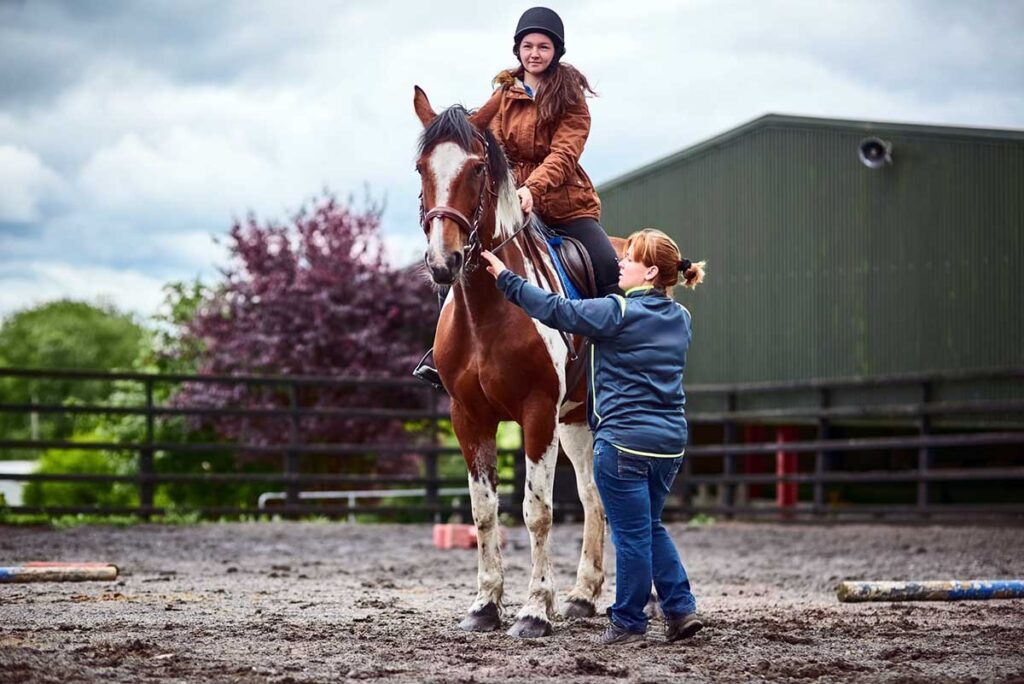 A horseback riding instructor teaches a novice rider on a brown and white paint horse her riding aids.