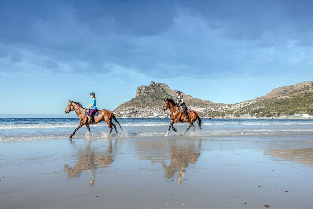 two young women gallop bay horses across a beach with mountains in the background during a horseback riding vacation