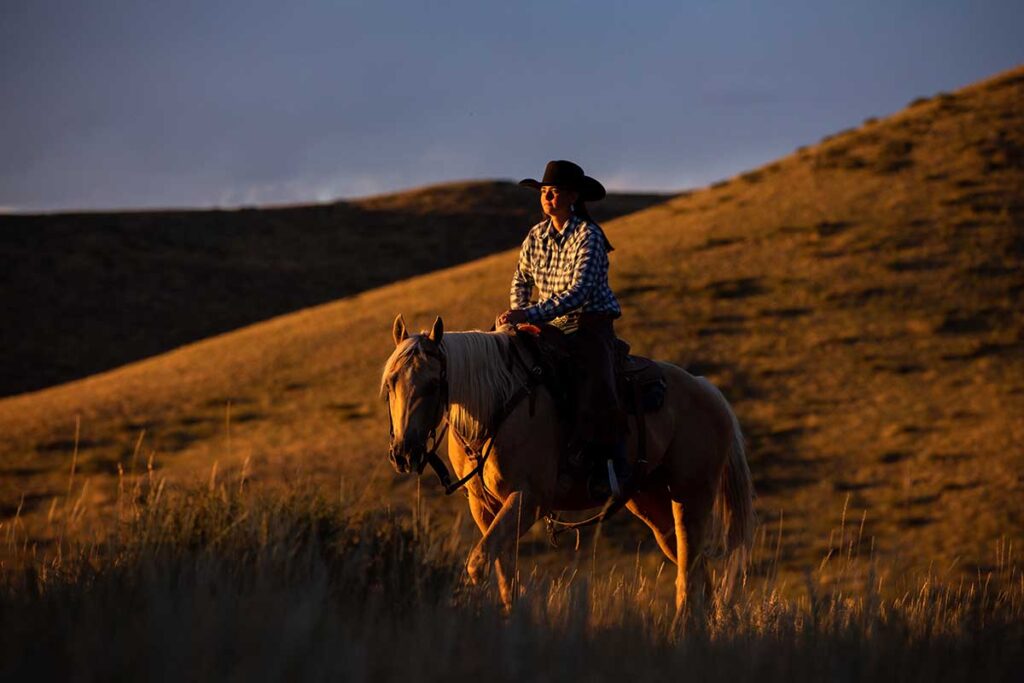 A cowgirl rides her palomino horse through the hills at sunset