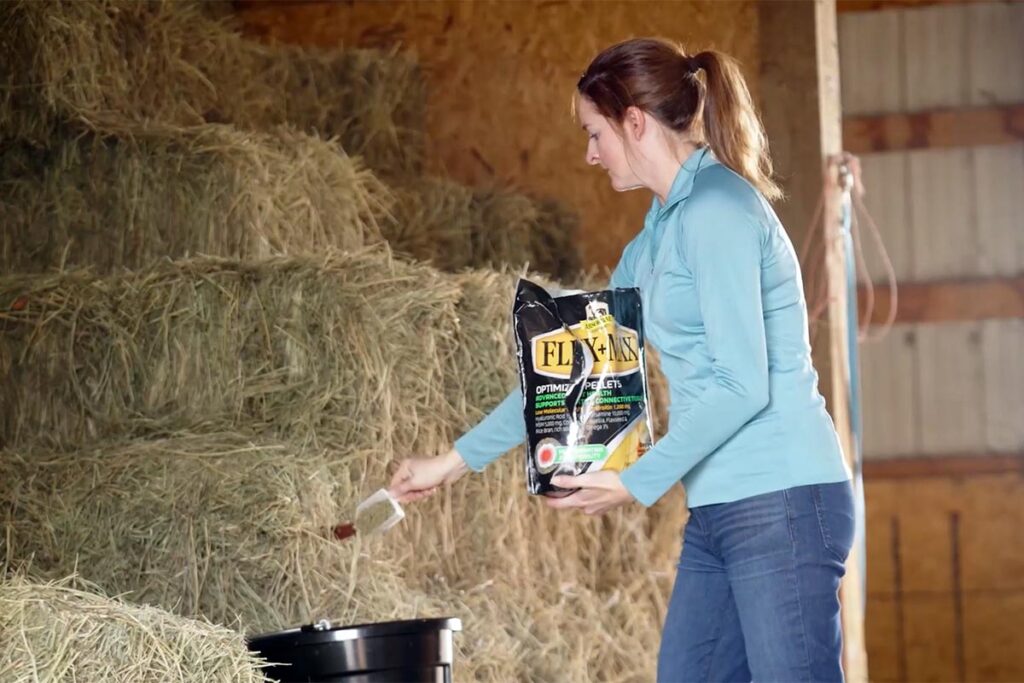 a woman adds a scoop of joint supplements for horses into a black bucket in a barn full of hay