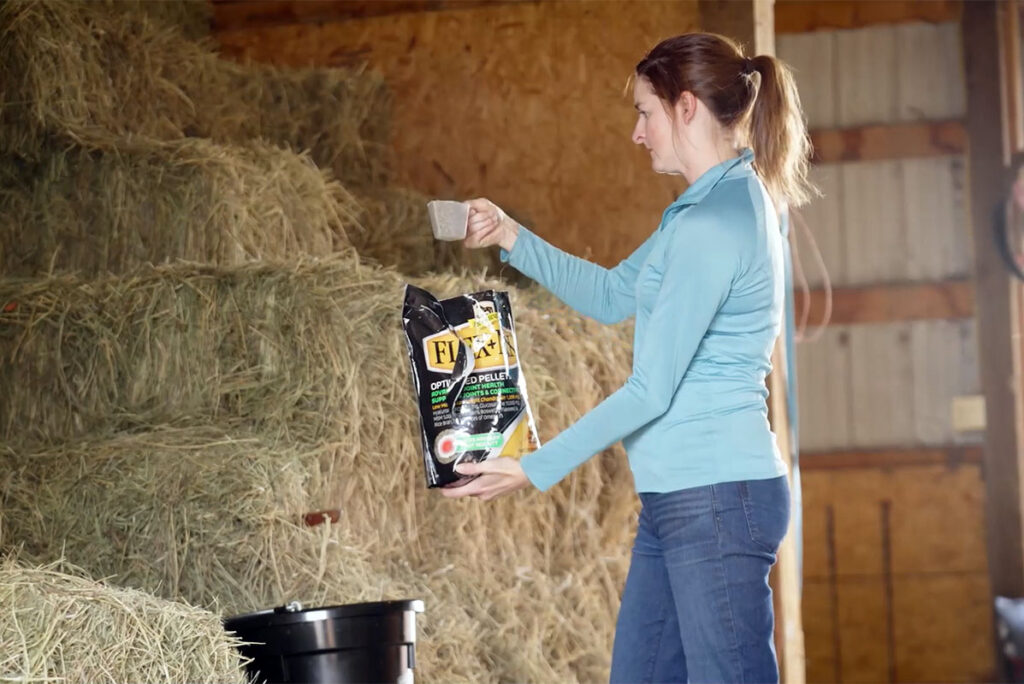 a woman measures out a scoop of a joint supplement for her horse before putting it in his feed bucket