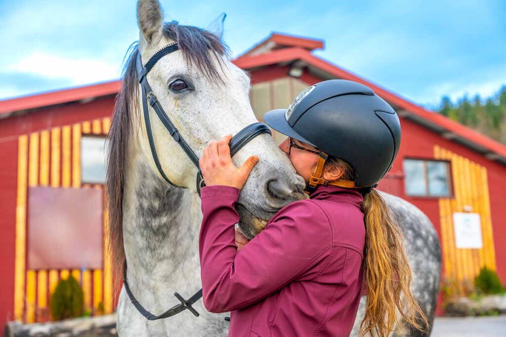 A girl wearing a riding helmet and English riding attire kisses her dapple gray horse on the nose after finding a horse that meets her needs perfectly.