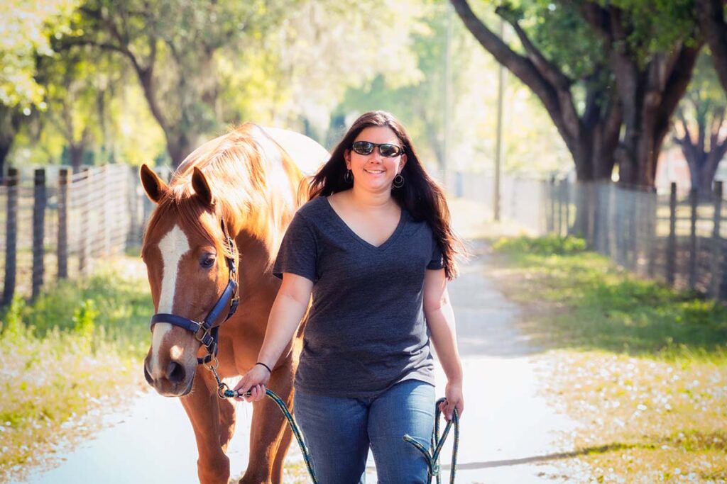 a woman wearing sunglasses in the summer leads her chestnut horse down a path and wonders how to stay cool at the barn