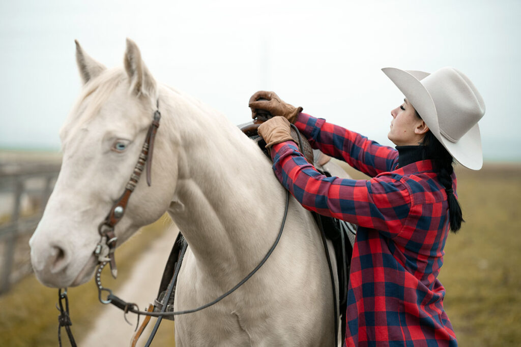a woman in western attire tacks up her cremello horse with a western saddle and checks its position on his back.