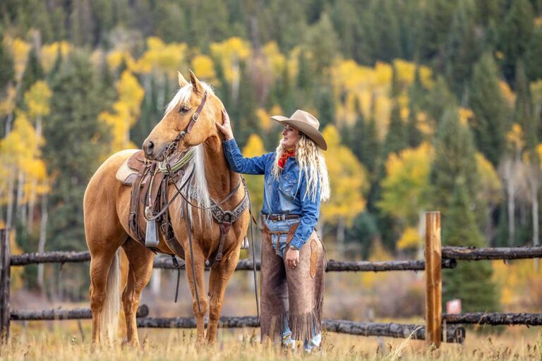 A woman in western gear and chaps stands next to her palomino horse while wondering what to pack for a fall horseback ride