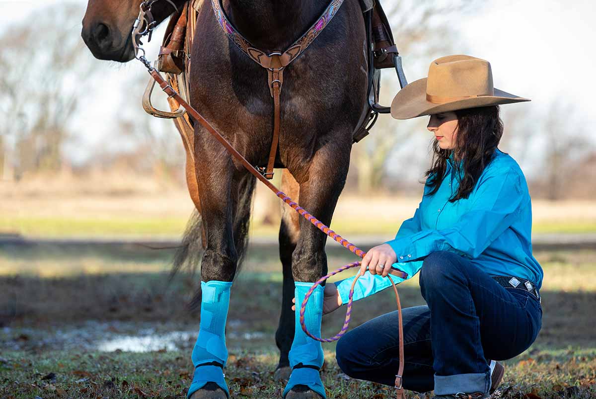 a cowgirl checks her bay horse's front legs while he wears blue boots.