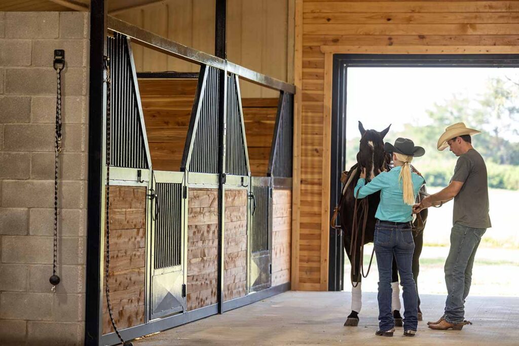 A man in a cowboy hat helps a cowgirl saddle her western horse in a barn aisle
