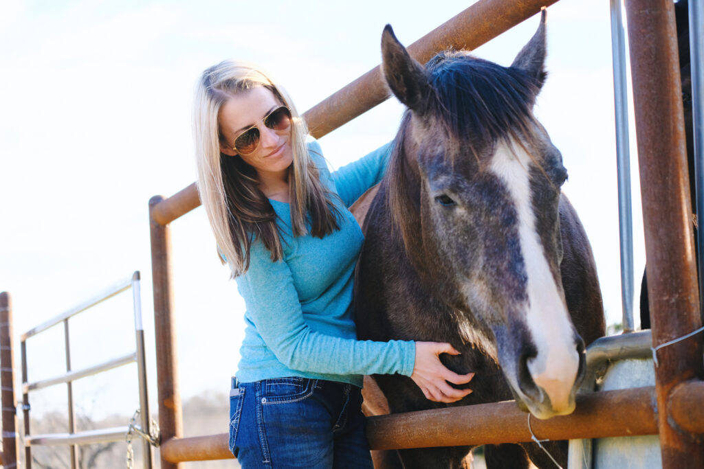 a woman in a blue shirt pets her older gray horse in a paddock