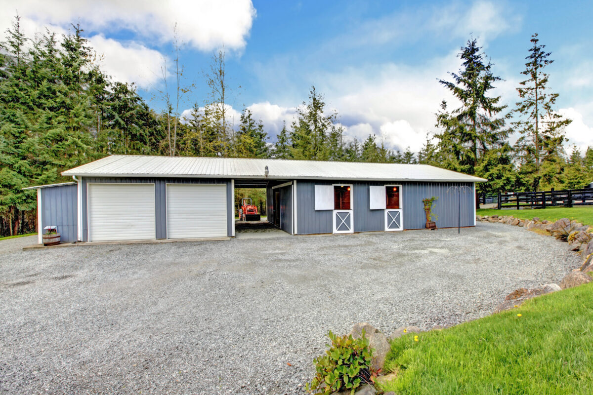 a small gray horse barn with two garage storage areas and a tractor parked behind it.